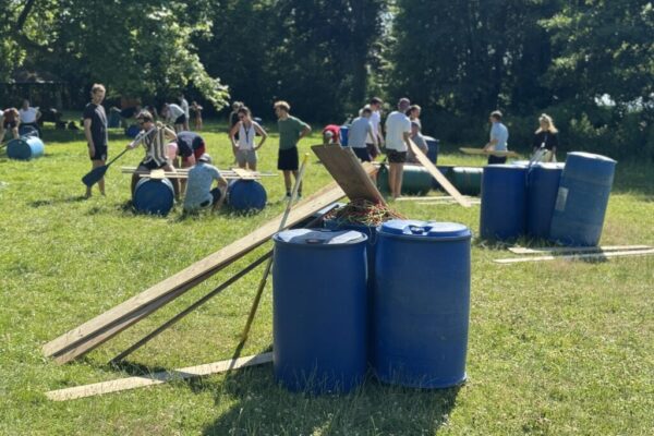 Auf einer grünen Wiese steht Baumaterial wie Plastiktonnen und Holz für den Floßbau.