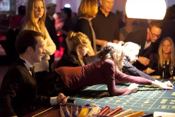 Mehrere Personen sitzen oder stehen um einen Roulette-Tisch herum. Eine blonde Frau lehnt sich über den gesamten Tisch, um ihren Einsatz zu platzieren.