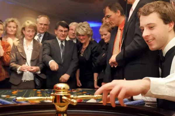 Bei einem Casinoabend sieht man im Vordergrund den Groupier, der das Roulette-Rad dreht, während die Zuschauer im Hintergrund gespannt das Ergebnis erwarten.